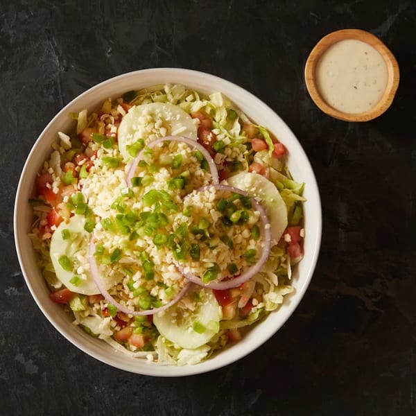 A large salad with lettuce, tomatoes, cucumbers, onions, cheese, and green peppers, with dressing on the side.