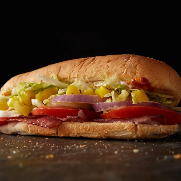A Plaza Pizza sub sandwich with salami, tomatoes, onions, lettuce, and peppers on a dark surface.