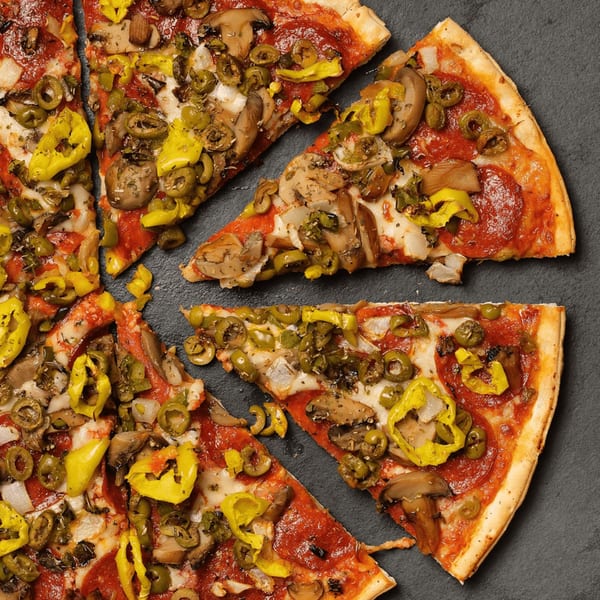 Plaza Pizza's pepperoni pizza with mushrooms, olives, and banana peppers, sliced on a dark surface.
