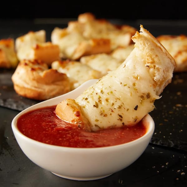 A cheesy garlic knot, seasoned with herbs, being dipped into a bowl of red marinara sauce.
