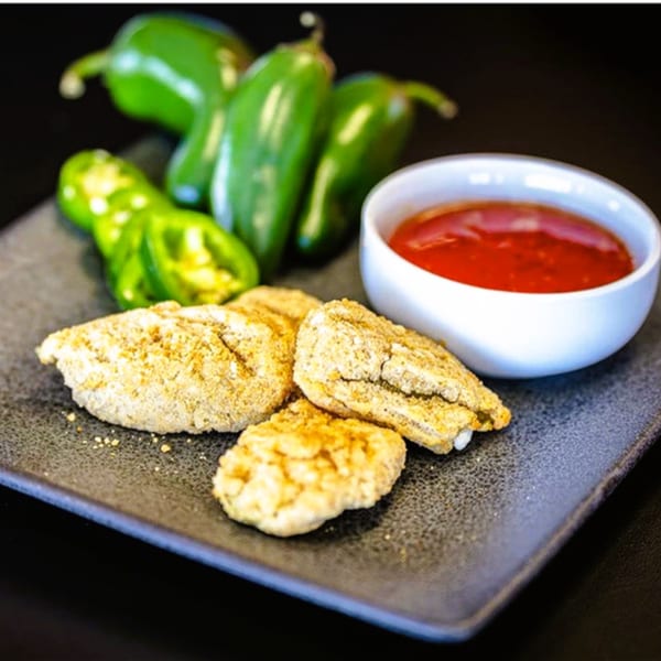 Breaded jalapeno poppers on a dark plate with a side of red dipping sauce and fresh green peppers.